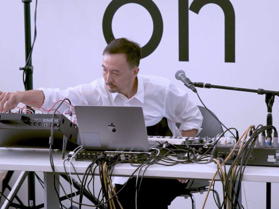 A masculine person sits at a table filled with recording and electronic equipment