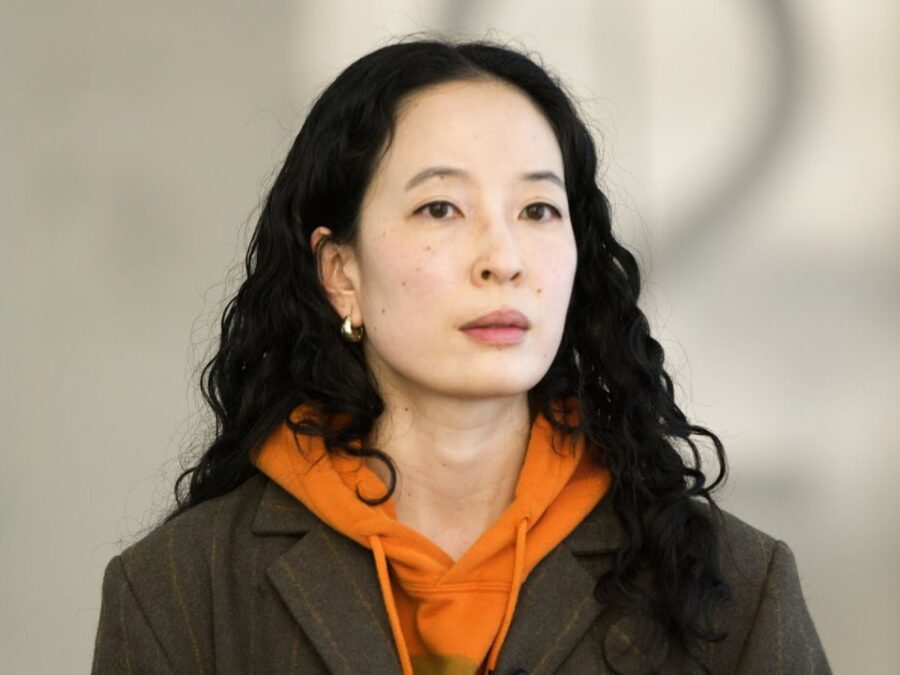 A female-presenting person with porcelain skin and dark brown curly hair in an orange hooded sweatshirt and brown blazer