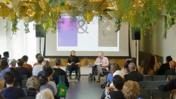 View from the back of an audience watching two people in front of them present something from the screen behind them in a room filled with hanging planters.