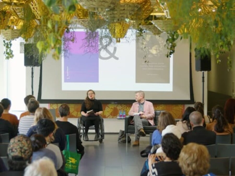 View from the back of an audience watching two people in front of them present something from the screen behind them in a room filled with hanging planters.