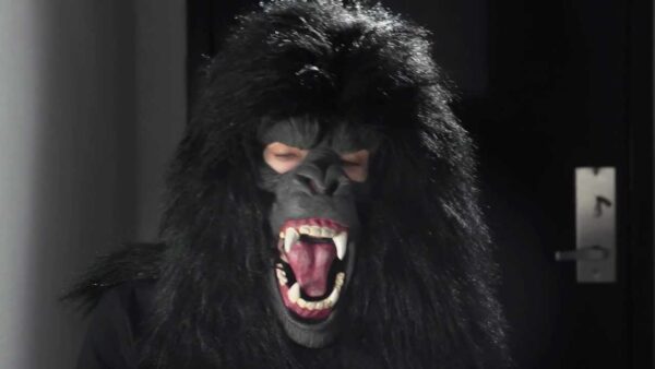 A person with a gorilla mask on sits in a darkened room