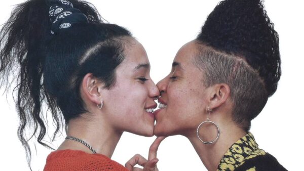 Two female presenting people share a kiss against a white background