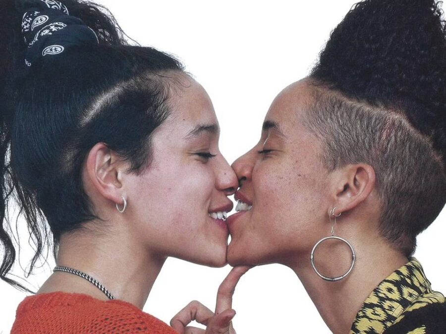 Two female presenting people share a kiss against a white background