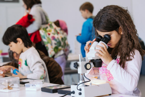 In a room filled with other children the focus is on a child with long brown hair looking through a microscope