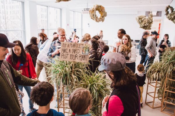 A group of people gather around pedestals filled with plants as brown wreaths hang overhead. A cardboard sign over one plant reads Pick a plantlet, make a card, post your plant.