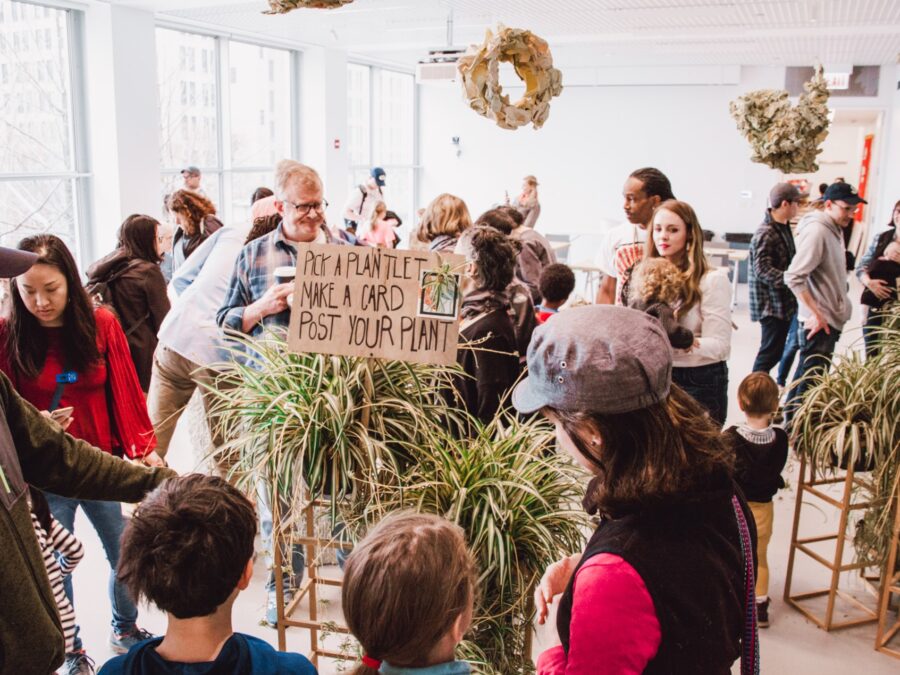 A group of people gather around pedestals filled with plants as brown wreaths hang overhead. A cardboard sign over one plant reads Pick a plantlet, make a card, post your plant.