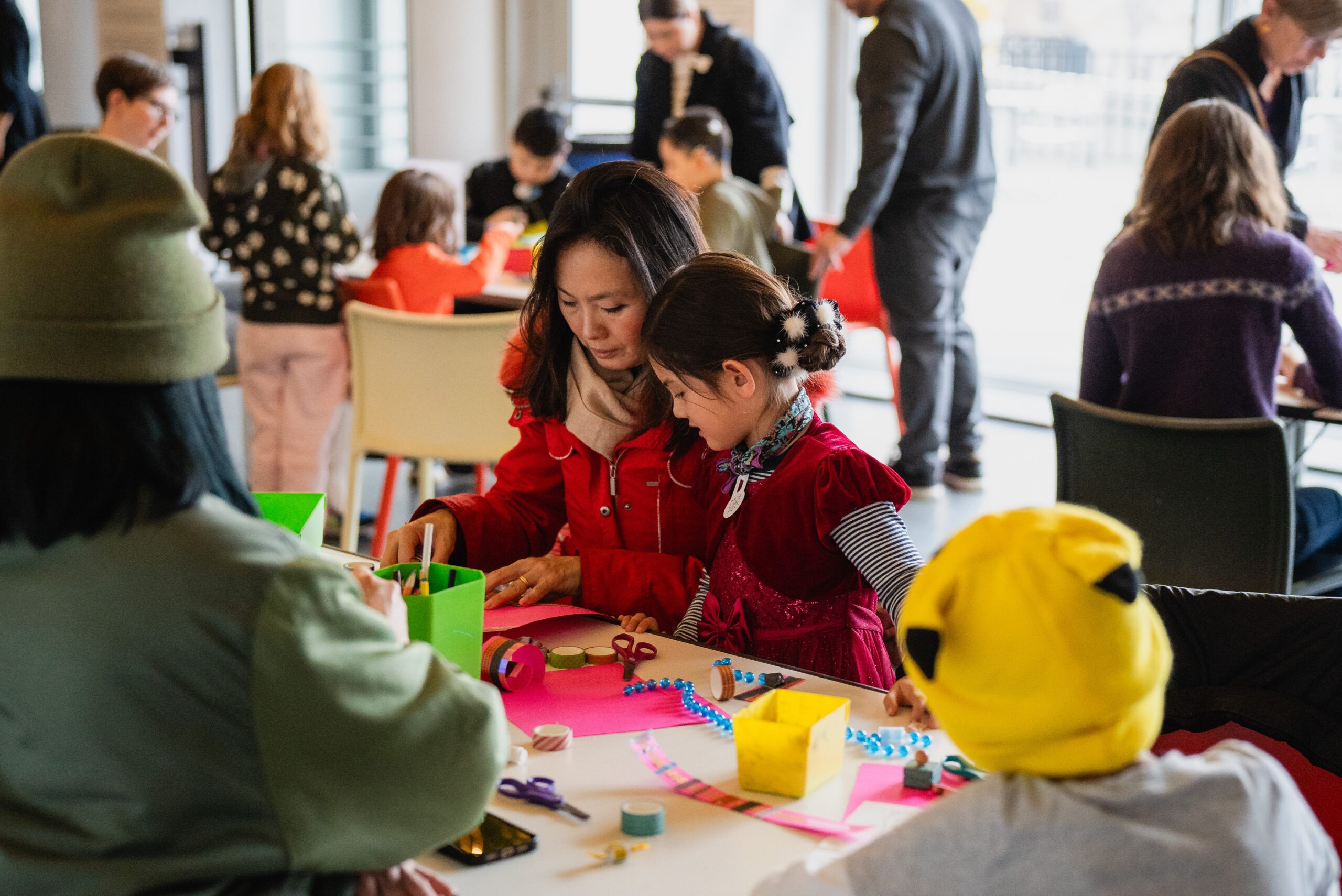 An adult and a child engaged in arts and crafts at a busy workshop table. Both are focused intently on their project which includes colorful papers and crafting materials.
