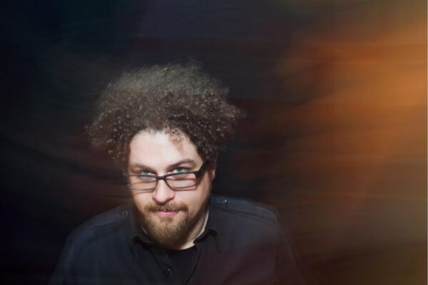 A male-presenting person with light skin and brown curly hair wearing glasses poses against a black background as an orange glare appears to their left