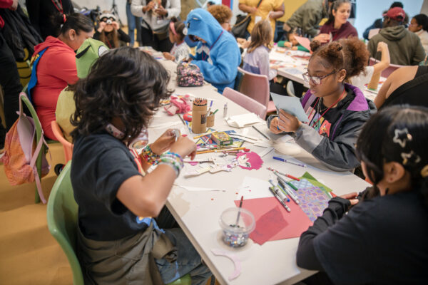 A group of people sitting around tables engaged in an arts and crafts activity. Various supplies like paper, markers, and scissors are scattered on the tables. Participants are focused on creating and decorating with colored paper and other materials. The environment is lively and collaborative.