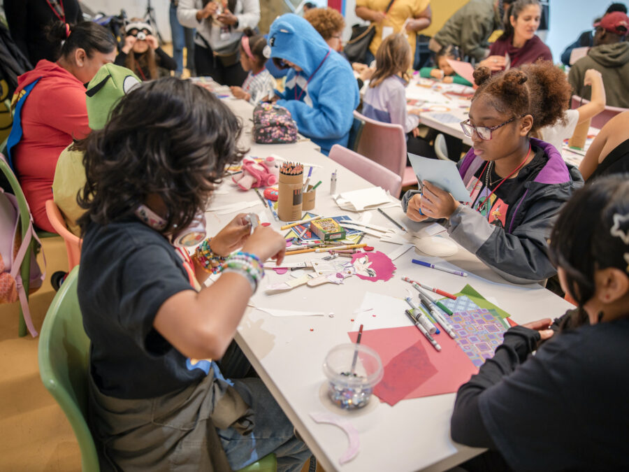 A group of people sitting around tables engaged in an arts and crafts activity. Various supplies like paper, markers, and scissors are scattered on the tables. Participants are focused on creating and decorating with colored paper and other materials. The environment is lively and collaborative.