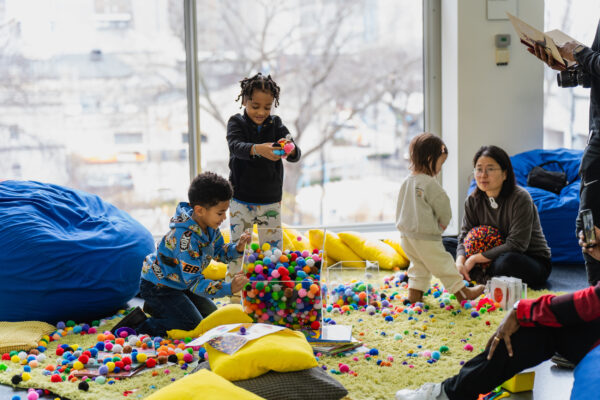 Children and adults play with an assortment of small puff balls from two clear containers on a yellow carpet surrounded by blue bean bag chairs and yellow pillows