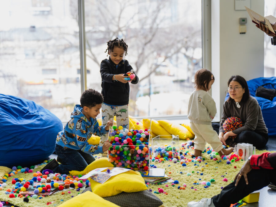 Children and adults play with an assortment of small puff balls from two clear containers on a yellow carpet surrounded by blue bean bag chairs and yellow pillows