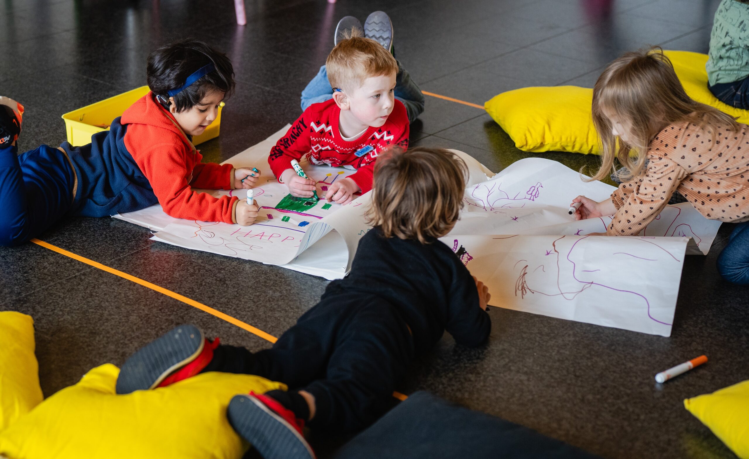 Four children color on a large paper on the floor surrounded by yellow pillows