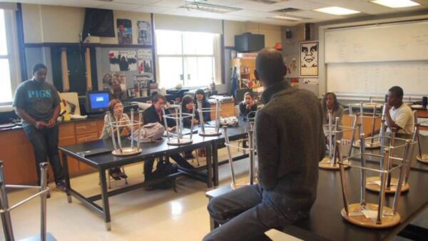 A person sits on a desk with their back to us in a classroom setting with students sitting at desks with stools placed upside down in front of them