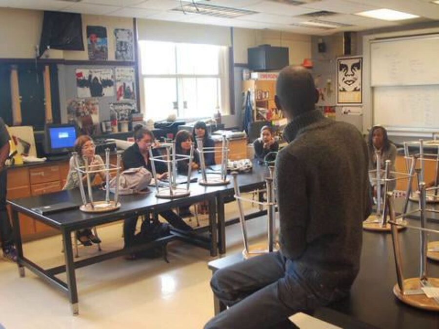 A person sits on a desk with their back to us in a classroom setting with students sitting at desks with stools placed upside down in front of them