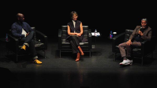 Three people sit on a black stage in black chairs. The person at stage left speaks into a microphone.