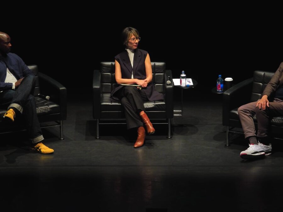 Three people sit on a black stage in black chairs. The person at stage left speaks into a microphone.