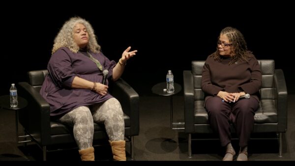 Two people sit in black chairs on a black stage. The person on the right leans in to listen to the person on the left.