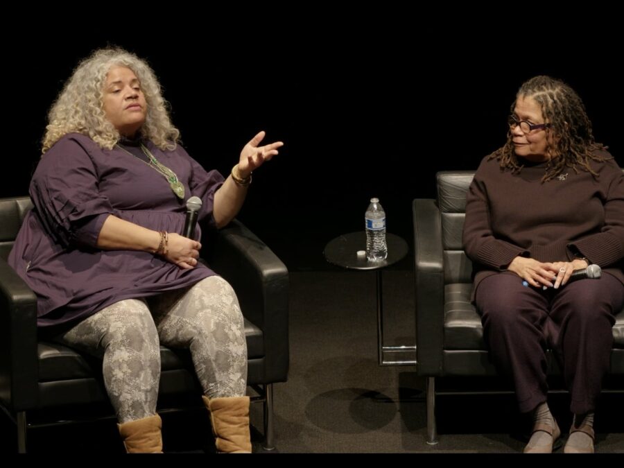 Two people sit in black chairs on a black stage. The person on the right leans in to listen to the person on the left.