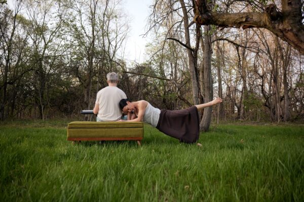 In a forest setting a person poses horizontally with their forearms resting on a green upholstered bench while extending one leg up next to another person sitting on the bench, their back to the viewer, with a keyboard on their knees