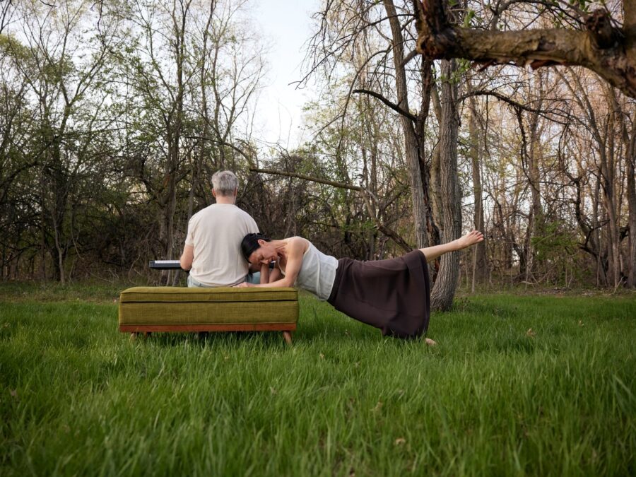 In a forest setting a person poses horizontally with their forearms resting on a green upholstered bench while extending one leg up next to another person sitting on the bench, their back to the viewer, with a keyboard on their knees