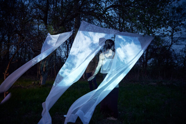 A person walks through white sheets hanging from a wire in a forest at dusk