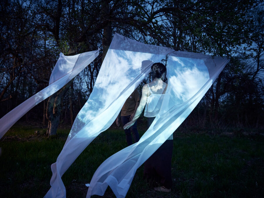 A person walks through white sheets hanging from a wire in a forest at dusk