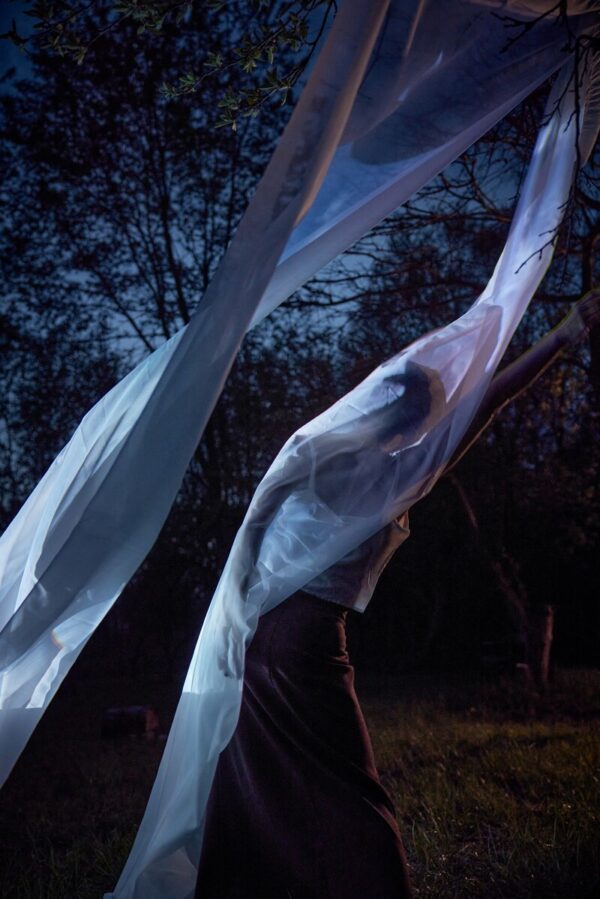 A person wrapped in a white sheet hanging from above in a forest at dusk
