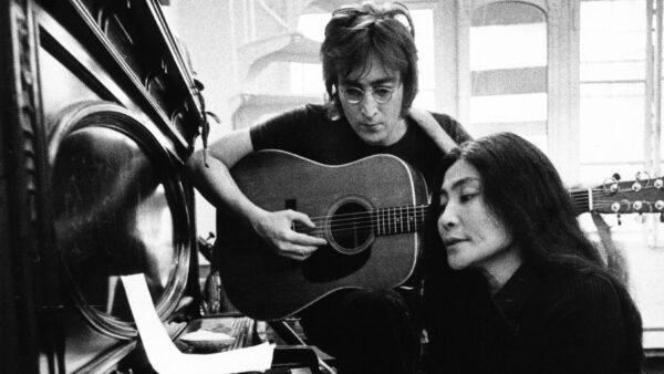 Black and white image of John Lennon playing an acoustic guitar and Yoko Ono reading lyrics at a piano in a bright room