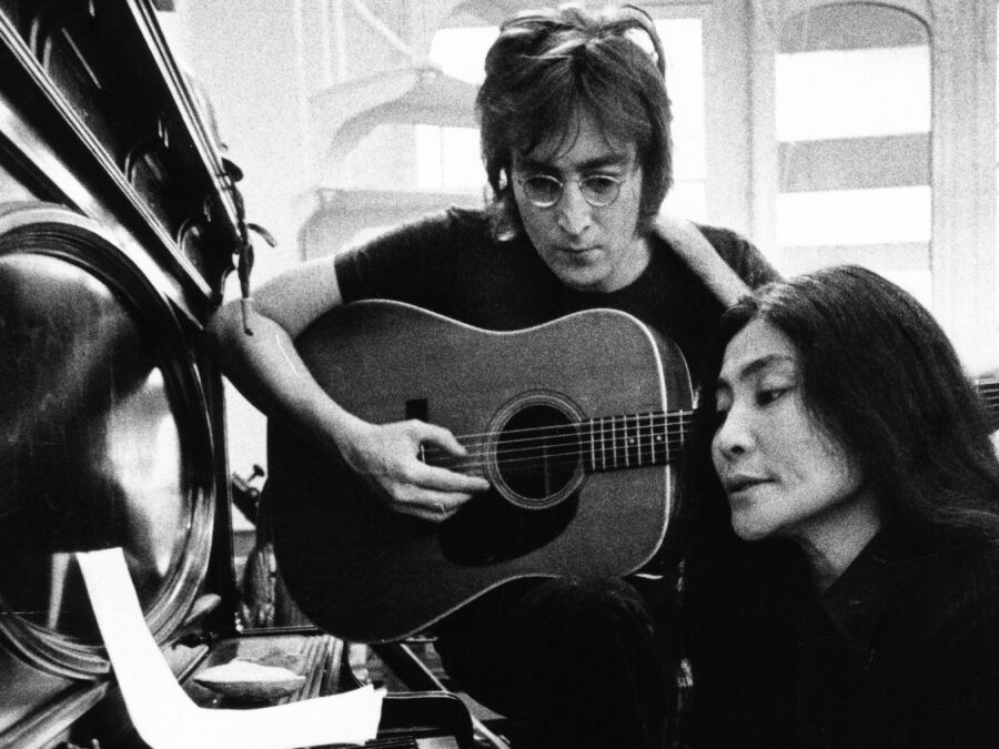 Black and white image of John Lennon playing an acoustic guitar and Yoko Ono reading lyrics at a piano in a bright room