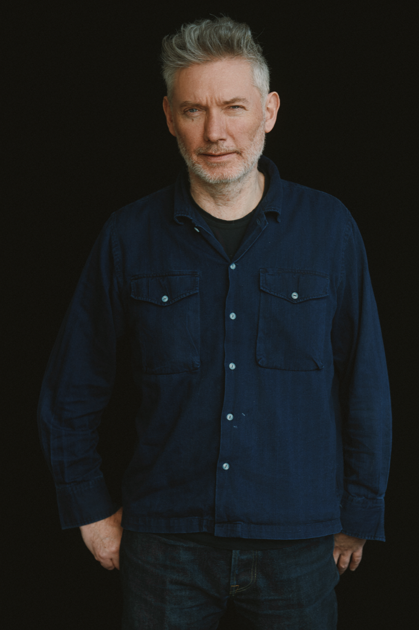Three-quarter portrait of a man with light skin and salt-and-pepper hair wearing a dark blue button down shirt against a black screen