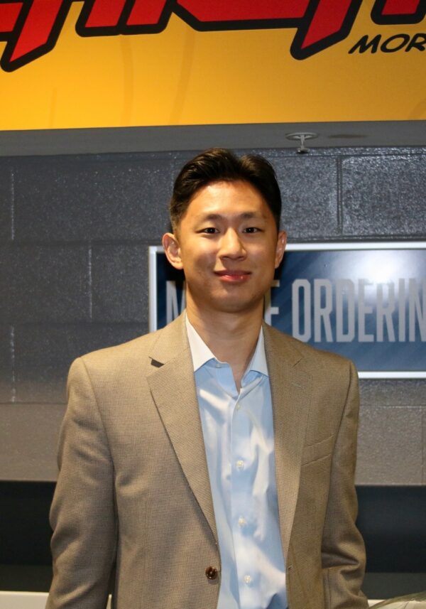A male-presenting person with medium skin and short brown hair in a tan suit coat and white shirt standing in front of a brick wall