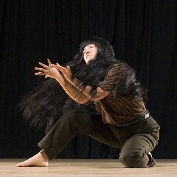 A person in midmotion their arms contorted in front of them as they kneel with one leg on the ground and the other bent to their right as they turn, their long black hair raised from the movement