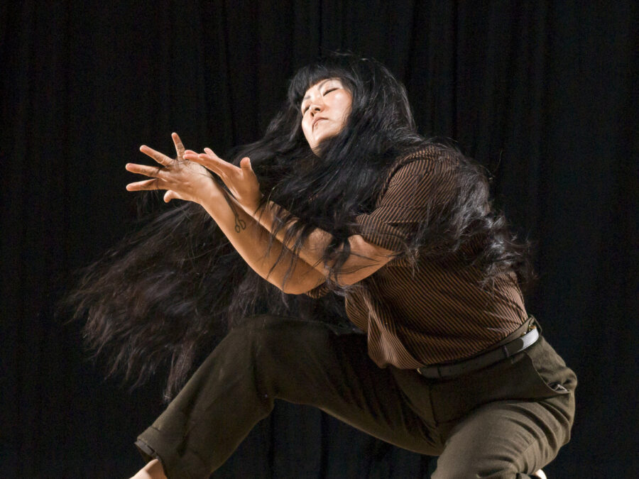 A person in midmotion their arms contorted in front of them as they kneel with one leg on the ground and the other bent to their right as they turn, their long black hair raised from the movement