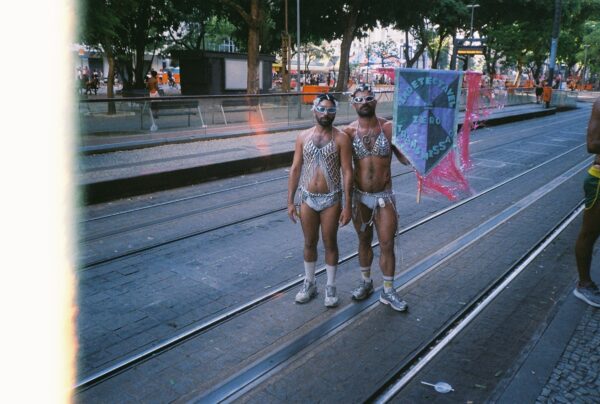 Two people in silver bikinis and glitter masks stand in the middle of tram tracks. The one on the right holds a banner reading Indetectavel = Zero Transmissao.