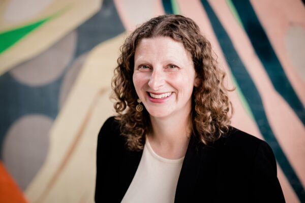 A female presenting person with light skin and light brown curly hair that's shoulder length smiles in front of an abstract mural