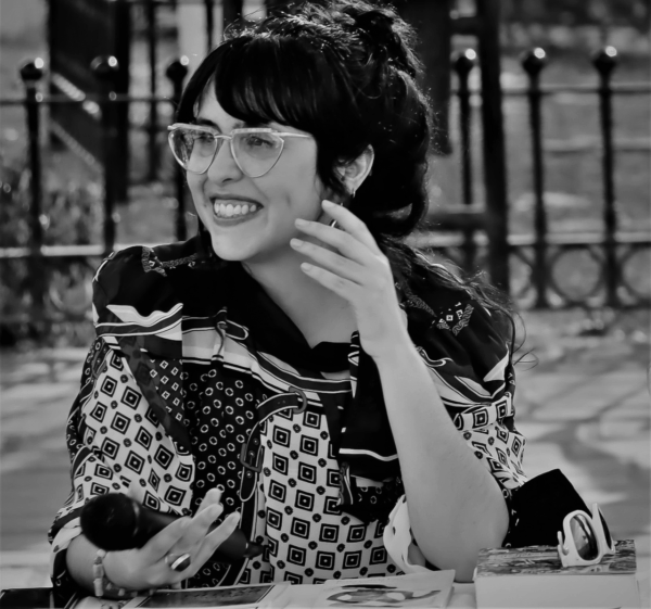 Black and white portrait of a female-presenting person with dark hair wearing a heavily patterned shirt. Their left hand is lifted to their jawbone as they look to their right