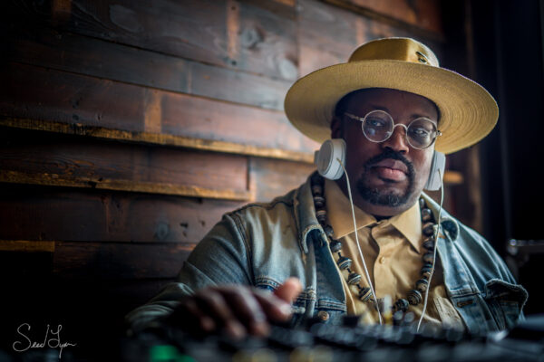 Portrait of a male-presenting person with dark skin, a salt-and-pepper goatee, and gold-rimmed glasses. They wear a wide-brimmed straw hat and headphones slightly on their ears.