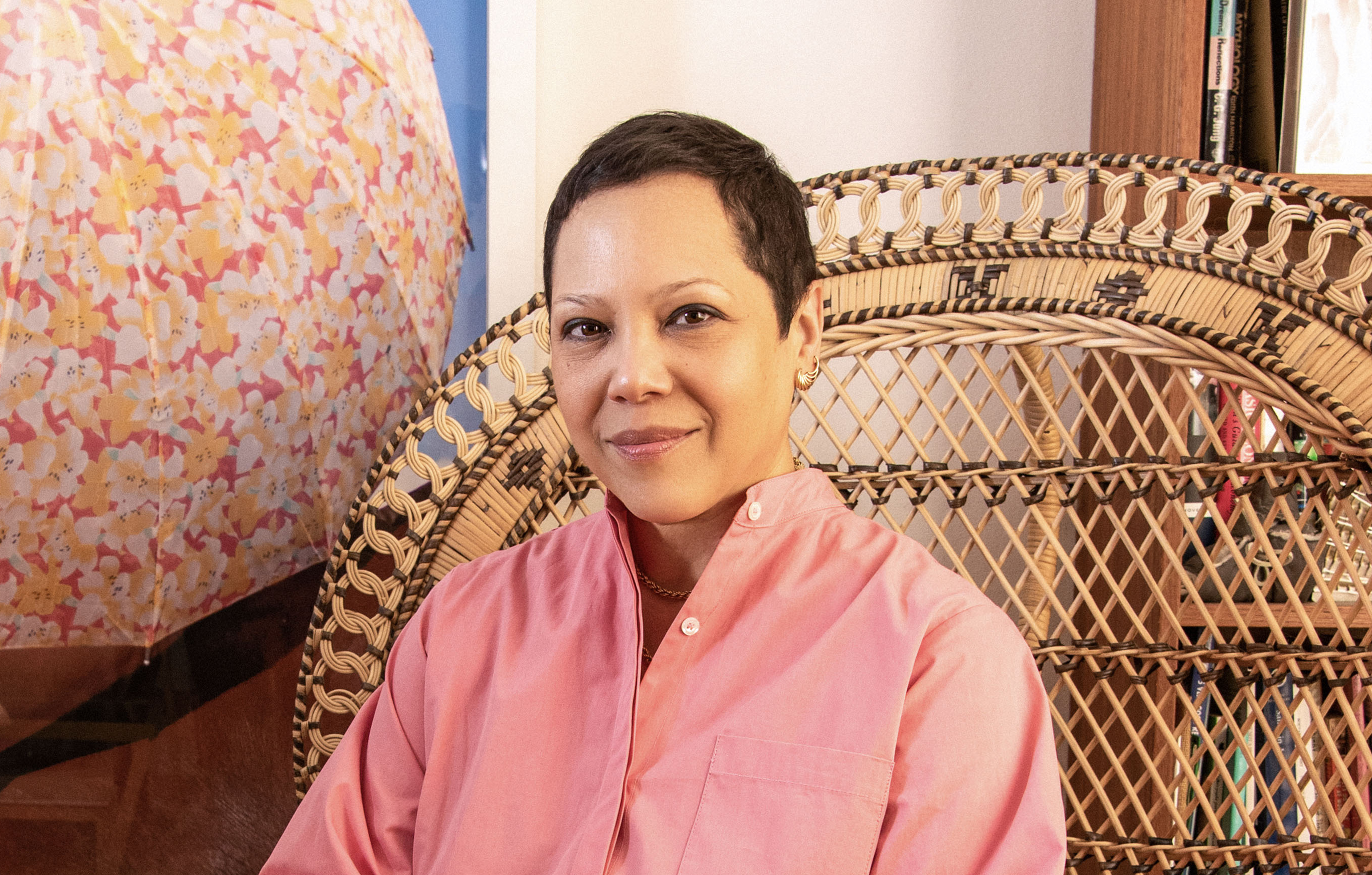 A female presenting person with medium tone skin and short brown hair poses in a peacock chair with a colorful, abstract artwork hanging to their right
