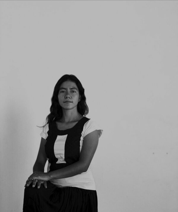 Black and white portrait of a female-presenting person posing against a solid light backdrop. They rest both hands on a raised knee.