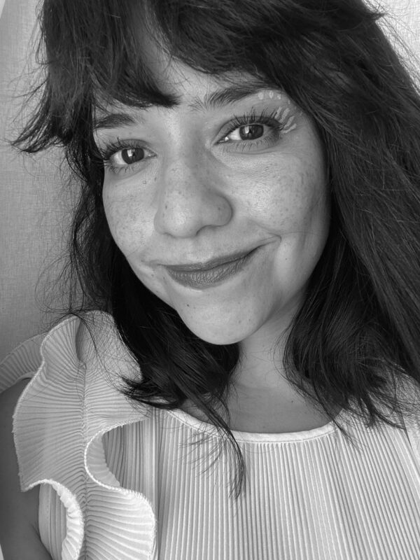 Black and white selfie of a female-presenting person with bangs and shoulder-length hair with a closed-lipped smile