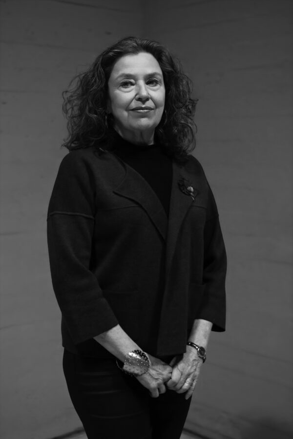 Black and white three-quarter portrait of a female-presenting person standing against a dark grey backdrop. They are dressed in all dark clothes and hold the pointer and middle finger of their right hand with their left hand at their waist.