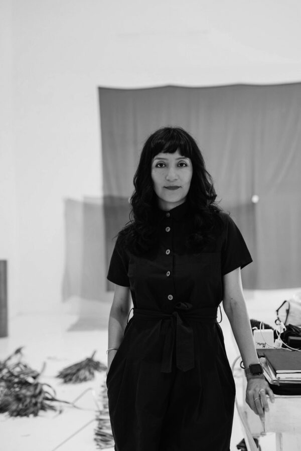 Portrait of a female person wearing a dark colored jumpsuit and standing in a studio space with fabric tacked to the walls
