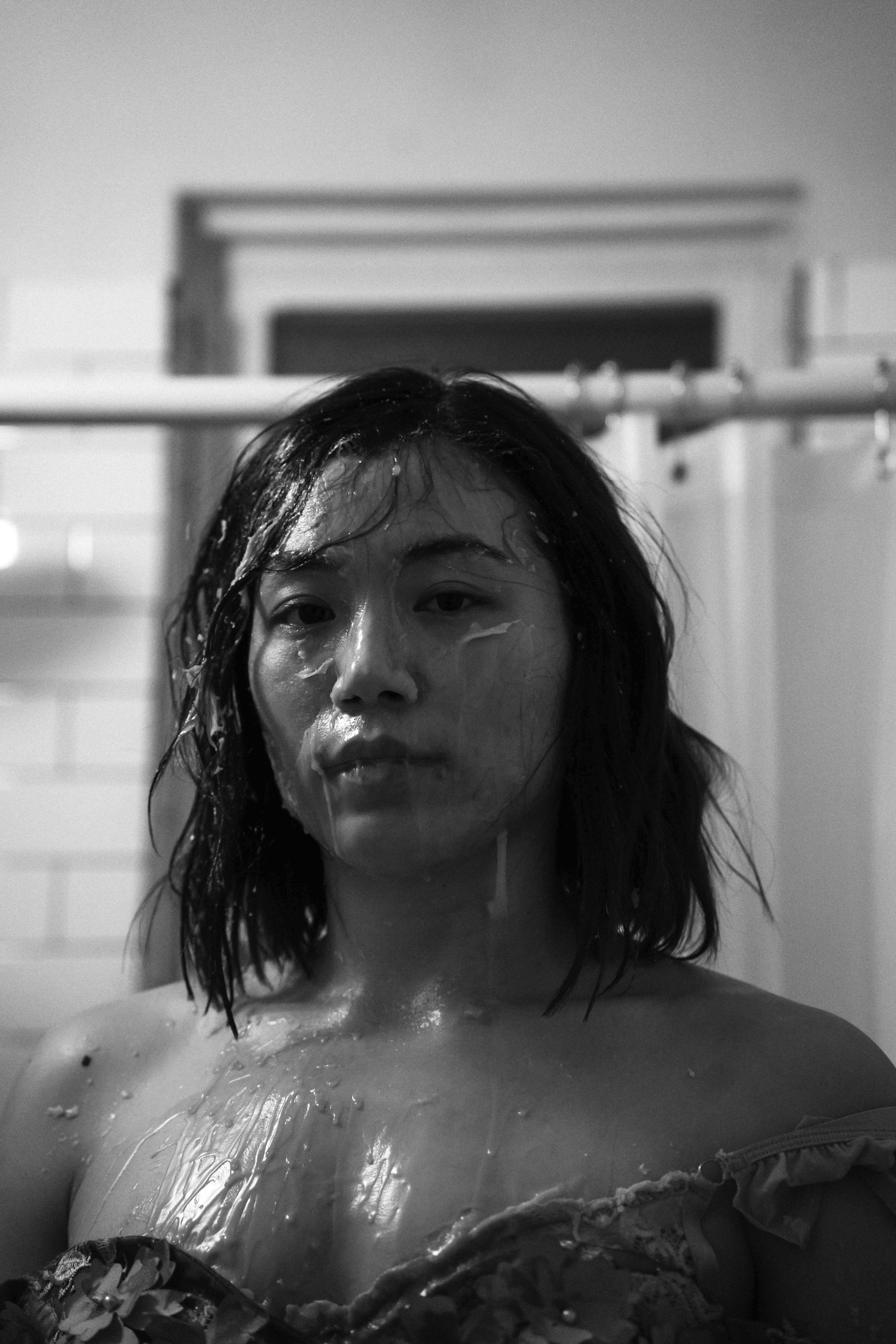 Black and white portrait of a female-presenting person with a shower curtain behind them and a liquid or gel spread across their face and chest