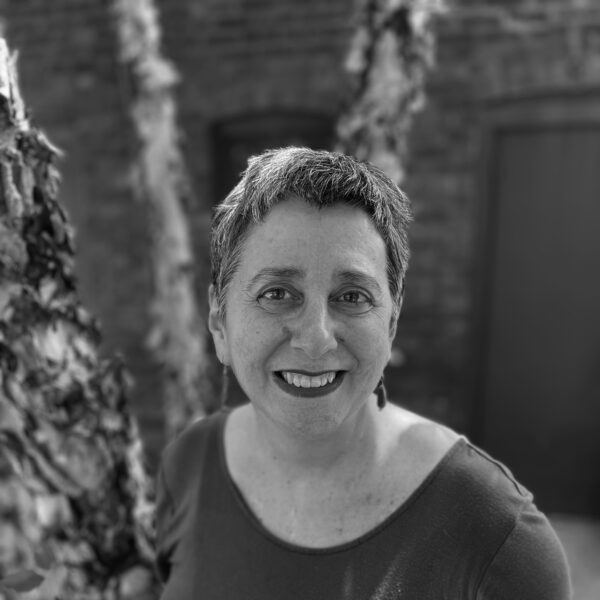 A black and white portrait of a female-presenting person with short, light hair posing outside near a tree