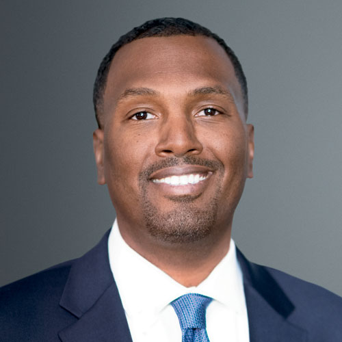 A male-presenting person with dark skin and short black hair smiles in a suit in front of a grey screen