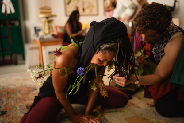 A person posing in frog position with the stems of wildflowers in their mouth as another person kneels next to them holding a spoon up to their face