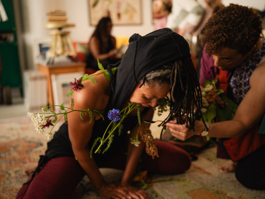 A person posing in frog position with the stems of wildflowers in their mouth as another person kneels next to them holding a spoon up to their face