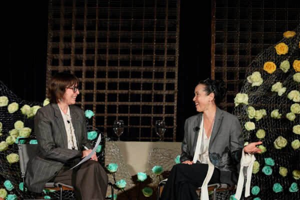 Two people sit on a stage in front of metal grids covered in teal, yellow, and pink flowers