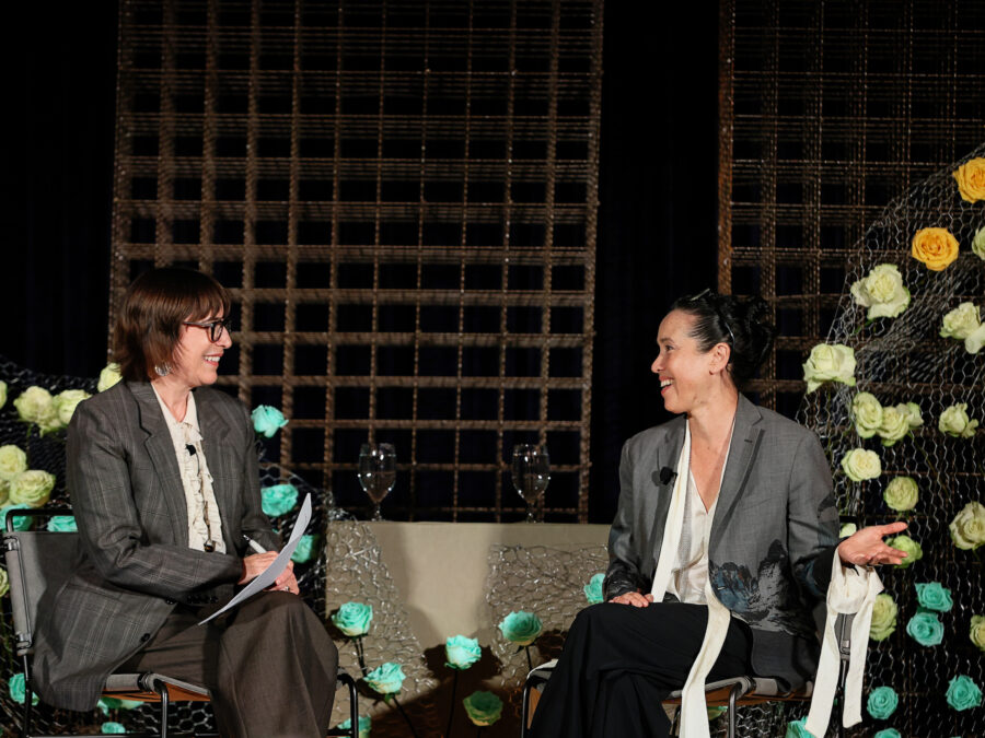 Two people sit on a stage in front of metal grids covered in teal, yellow, and pink flowers